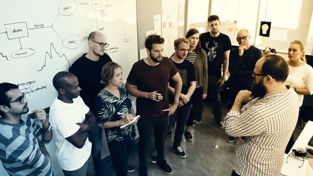 A team has a stand up meeting in front of a whiteboard.