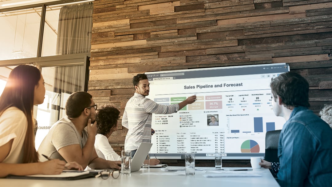 A man points at a Smartsheet dashboard on a large monitor during a meeting.